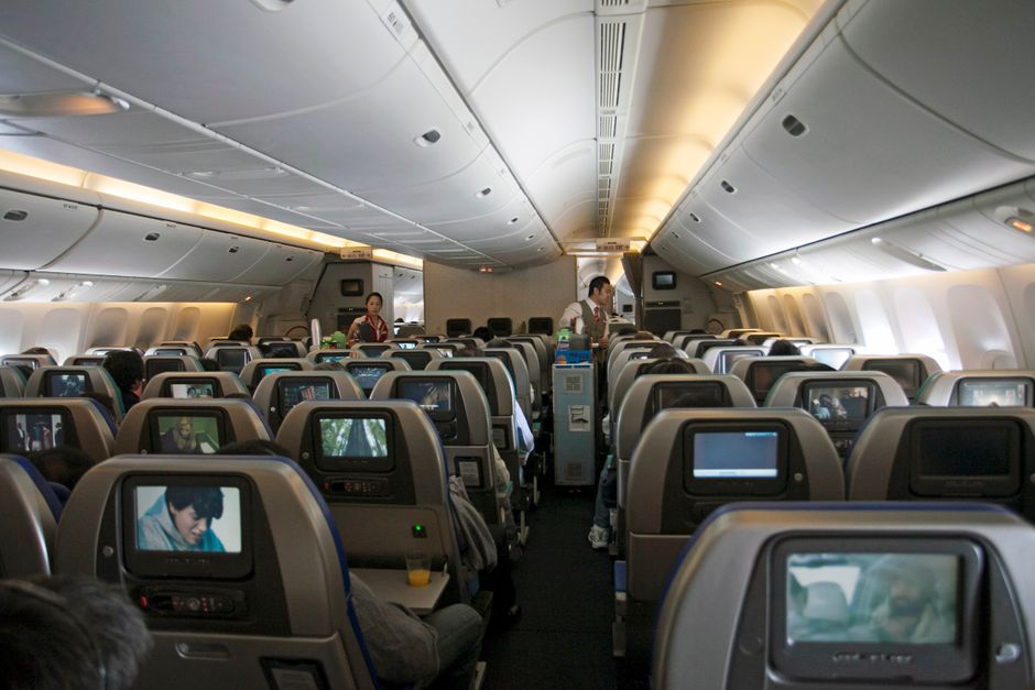 In-flight view of an aircraft cabin featuring passengers, flight attendants, and seatback screens.